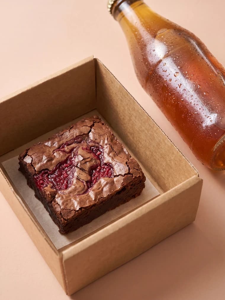 Close-up of a fudgy chocolate brownie in the corner of a kraft lunch box beside a chilled bottle of iced tea