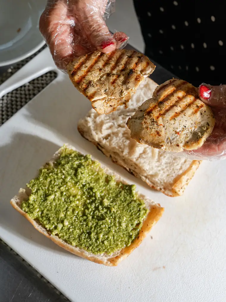 Grilled chicken being placed onto a pesto-spread ciabatta while a catering order is prepared