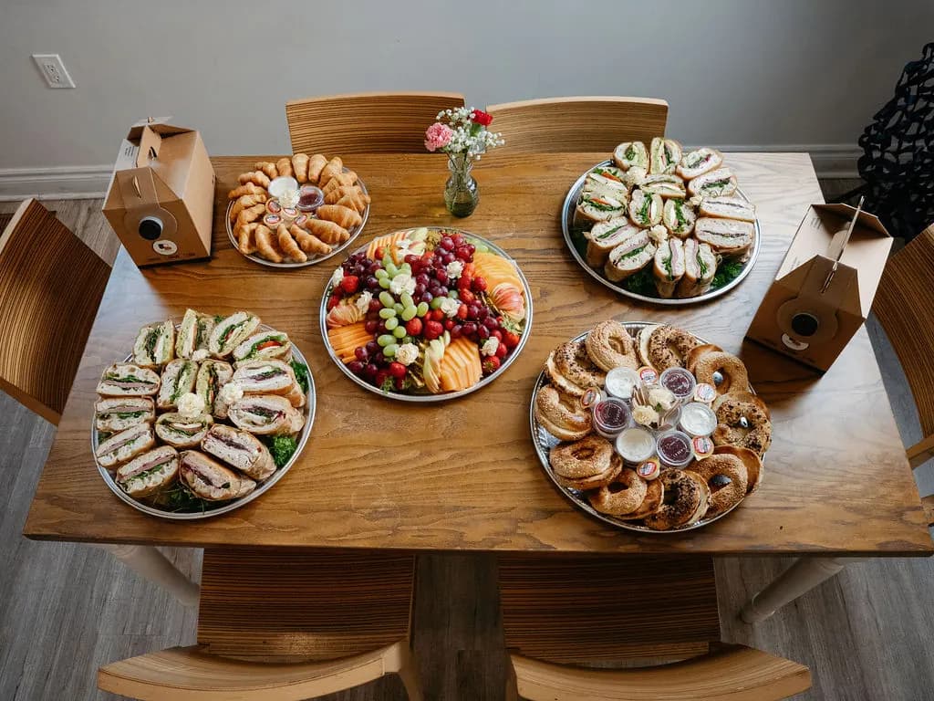 Overhead view of a catered office table with two sandwich trays, fruit, croissants, bagels and coffee boxes