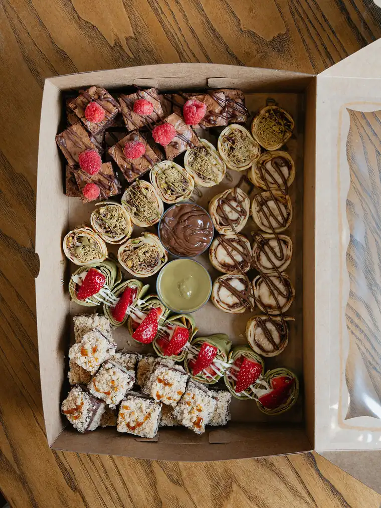 Dessert catering box with raspberry brownies, pistachio and Nutella crepe rolls, crumble bars and dipping sauces